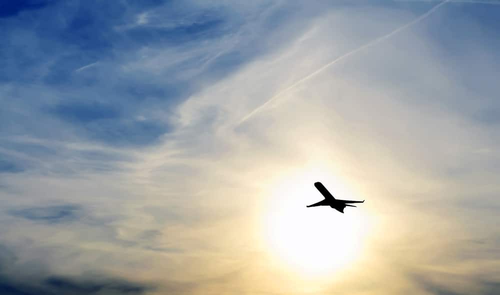 A silhouette of a jet against the sun in a cloudy blue sky.