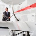 A businessman in a suit holding a briefcase looks back before climbing aboard a private jet with a red stripe down the fuselage.