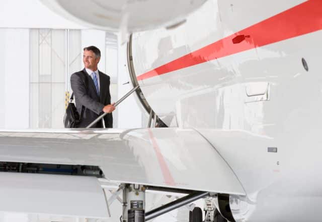 A businessman in a suit holding a briefcase looks back before climbing aboard a private jet with a red stripe down the fuselage.