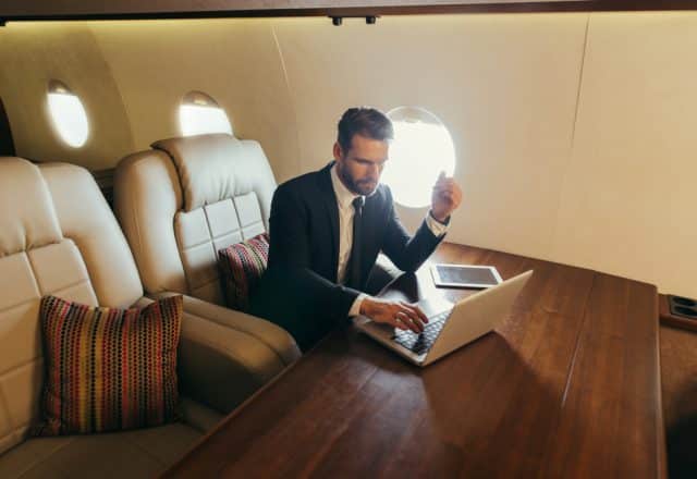 A business man in a suit works on his laptop at a table inside a private jet.