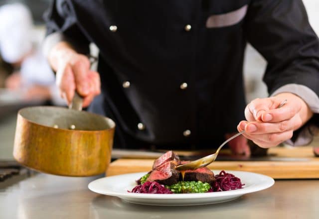 A chef plates a beautiful five-star meal, gently spooning sauce onto a delicious steak dish.