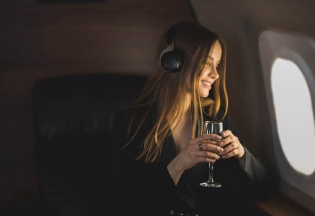 A woman wearing headhpones enjoys a glass of wine while looking out the window of her private jet.