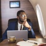 A man sits infront of a laptop at a desk inside a private jet.