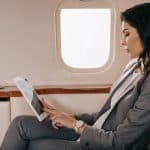 A woman in a grey pant suit works on a tablet while sitting comfortably with her legs crossed on a private jet.