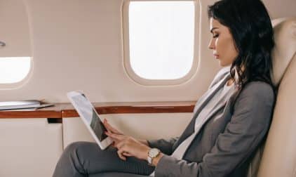 A woman in a grey pant suit works on a tablet while sitting comfortably with her legs crossed on a private jet.