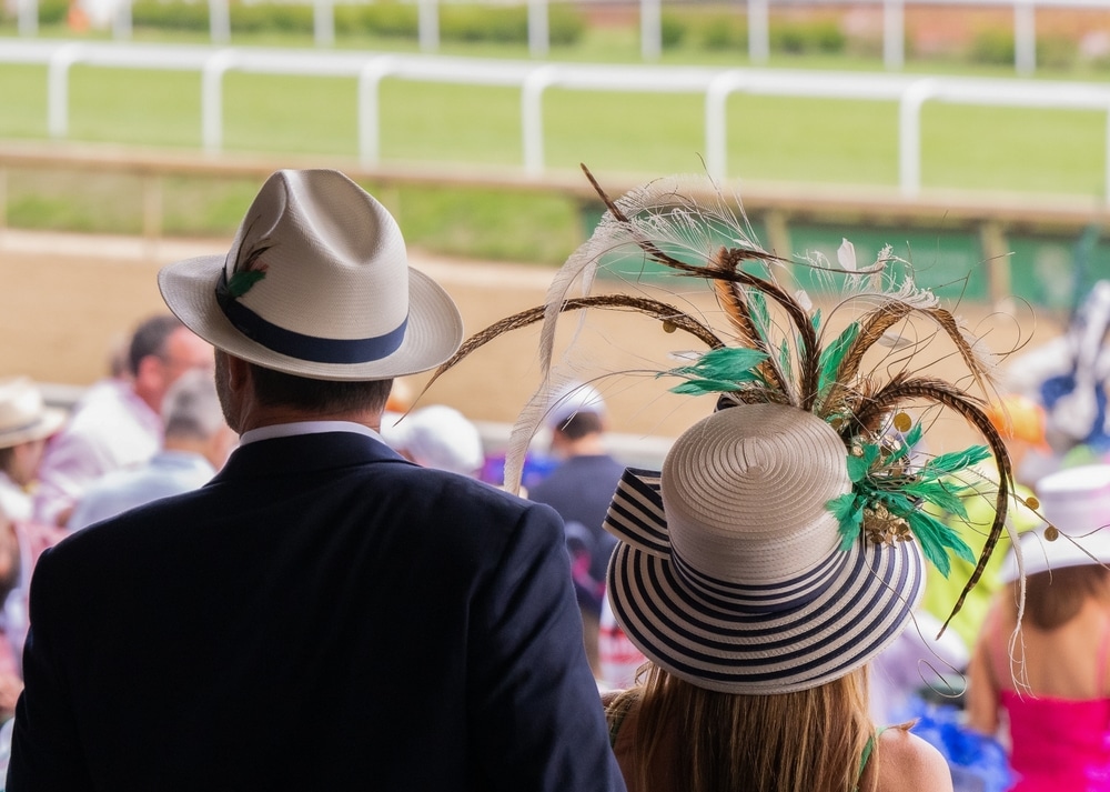 A man and a woman with elegant wide-brimmed hats sit in the stands to watch the Kentucky Derby.